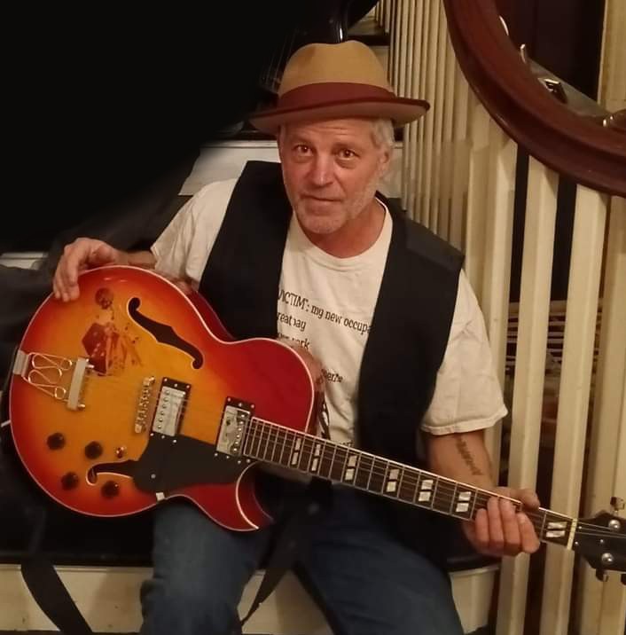 Stevie “C-Note” Cunningham smiling, holding a sunburst hollow-body electric guitar